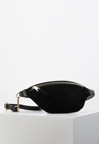 Riñonera de ante negro con forma curvada, detalles en dorado en la cremallera y acentos de cuero negro. Correa ajustable para un uso versátil.