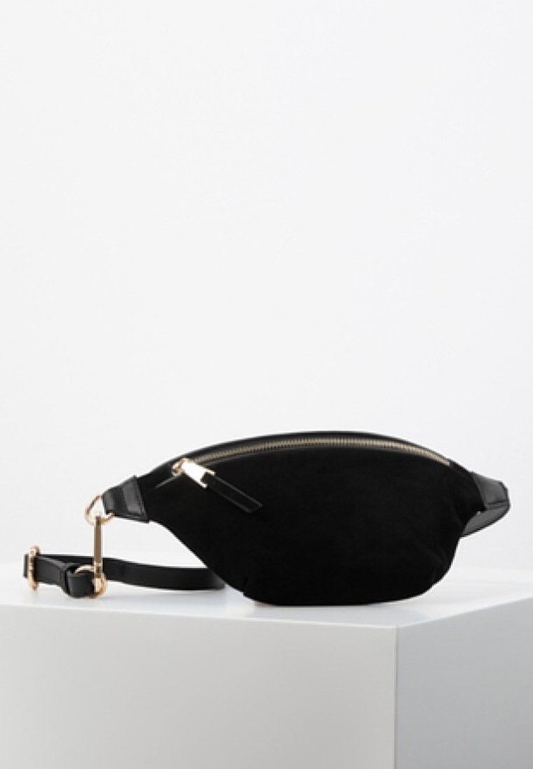 Riñonera de ante negro con forma curvada, detalles en dorado en la cremallera y acentos de cuero negro. Correa ajustable para un uso versátil.