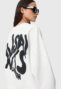 Felpa bianca oversize con un grande design di testo astratto nero sulla schiena; modello che indossa occhiali da sole neri angolari.