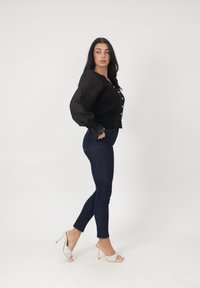 Mujer de perfil vistiendo una blusa negra, jeans ajustados oscuros y zapatos blancos de tacón alto y con punta abierta frente a un fondo liso.
