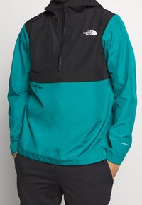 Giacca pullover nera e teal, con zip frontale a metà, cappuccio regolabile e dettaglio del logo sul petto e sulla manica. Materiale leggero.