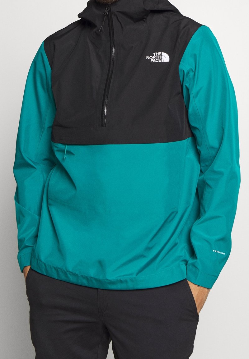 Giacca pullover nera e teal, con zip frontale a metà, cappuccio regolabile e dettaglio del logo sul petto e sulla manica. Materiale leggero.