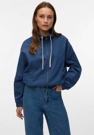 Donna che indossa una giacca con zip blu con coulisse bianche e jeans blu a vita alta, in piedi davanti a uno sfondo bianco uniforme.