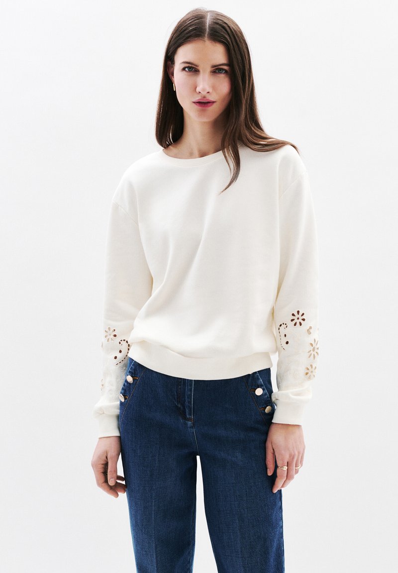 Felpa crema con ricami floreali intagliati sulle maniche, abbinata a pantaloni in denim blu con dettagli in bottone dorato.