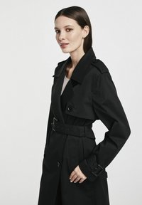 Czarny płaszcz trench z paskiem w talii, dużymi klapami i zapinany na guziki. Posiada długie rękawy i gładki, strukturalny materiał.