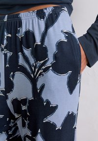 Personne portant un pantalon bleu clair avec de grands motifs floraux noirs, une main dans la poche latérale, et une chemise longue à manches sombres partiellement visible.