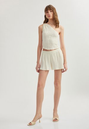Top crema de un solo hombro con un diseño texturizado y fruncido. Combinado con pantalones cortos holgados a juego. Llevado con sandalias de tacón beige.