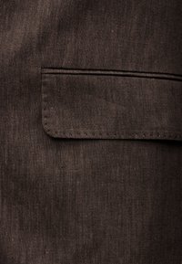 Nærbilde av mørkebrunt stoff med en sydd rektangulær klaffelomme på overflaten.