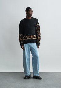 Fekete kötött pulóver színes mintás ujjakkal, világoskék, bővödő farmernadrággal és fekete cipővel párosítva. Egyszerű, kerek nyakkivágás.