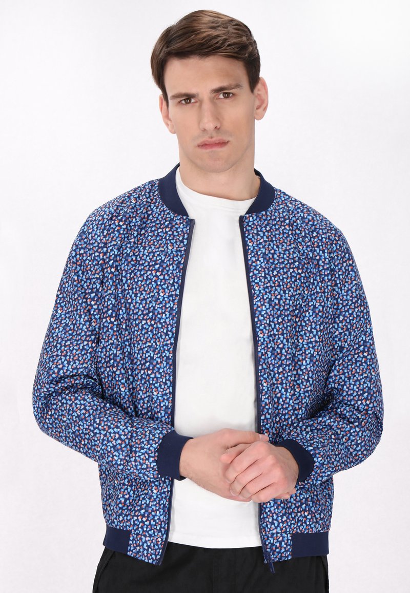 Marine bomjasje met een geometrisch patroon in blauw, rood en wit. Heeft een geribde kraag en manchetten. Draagt over een wit t-shirt.