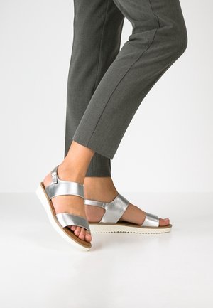 Sølvfarvede lædersandaler med justerbare stropper, en hvid gummisål og en glat tekstur, der viser åbne tæer og et moderne design.