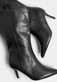 Mustat nahkaiset polven yli ulottuvat saappaat, joissa on terävä kärki, ompeluyksityiskohtia ja ohut korko. Liukas pinta, tyylikäs muotoilu.