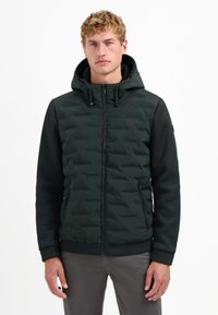 Man in een donkergroene gewatteerde hoodie met gestructureerde mouwen, met de rits dicht, staande tegen een witte achtergrond.