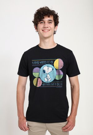 Giovane uomo sorridente, indossa una maglietta nera con illustrazione di Snoopy e testo "Non ho mai voluto essere altro che un cane" con motivi circolari colorati.