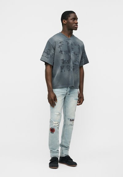 Άνδρας που φοράει ξεθωριασμένη μαύρη φαρδιά αθλητική φανέλα με τον αριθμό 99 και ανοιχτό μπλε σκισμένο τζιν με patches, στέκεται μπροστά σε απλό φόντο.