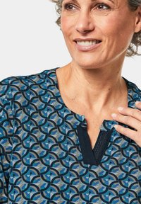 Musterbluse mit einem blauen und grauen geometrischen Design, einem dunkelblauen Kragen und einem einzigartigen Ausschnitt. Kurzärmlig, aus weichem Stoff.