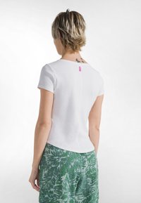 Bílý krátký rukávový top s vroubkovanou texturou a růžovou etiketu na zadní straně; doplněn zelenými kalhotami s potiskem.