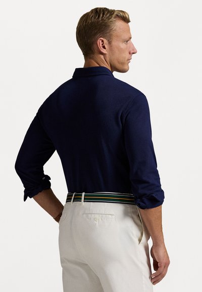 Navy blue long-sleeve polo shirt with rolled cuffs, paired with cream-colored pants and a multicolored striped belt.