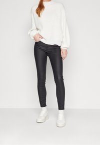 Mujer vestida con un suéter grueso blanco, pantalones ajustados negros, calcetines blancos y zapatillas blancas, de pie contra un fondo neutro.