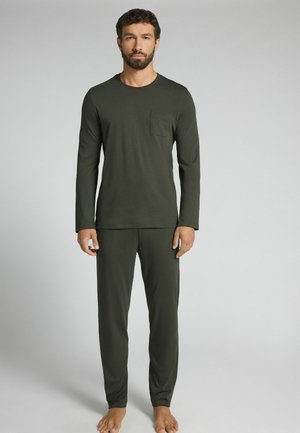 Man staat op blote voeten, draagt een donkergroene longsleeve met borstzak en bijpassende broek tegen een effen lichte achtergrond.