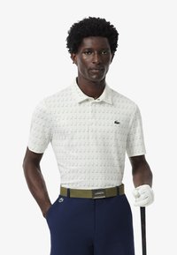 Polo a maniche corte di colore bianco con pattern verdi a tema golf. Accompagnato da una cintura verde oliva e pantaloni blu, indossato con un guanto da golf.