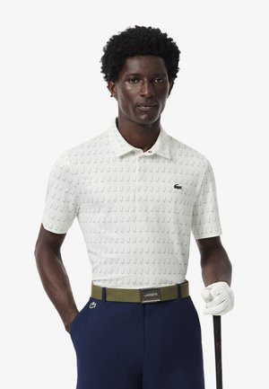 Polo a maniche corte di colore bianco con pattern verdi a tema golf. Accompagnato da una cintura verde oliva e pantaloni blu, indossato con un guanto da golf.