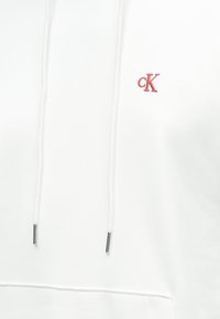 Sudadera blanca con capucha y logo "ck" bordado en rojo. Cuenta con cordones con terminales metálicos y una textura de tela suave.