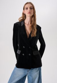 Vrouw met lang blond haar draagt een zwarte double-breasted fluwelen blazer en lichtblauwe spijkerbroek tegen een effen witte achtergrond.