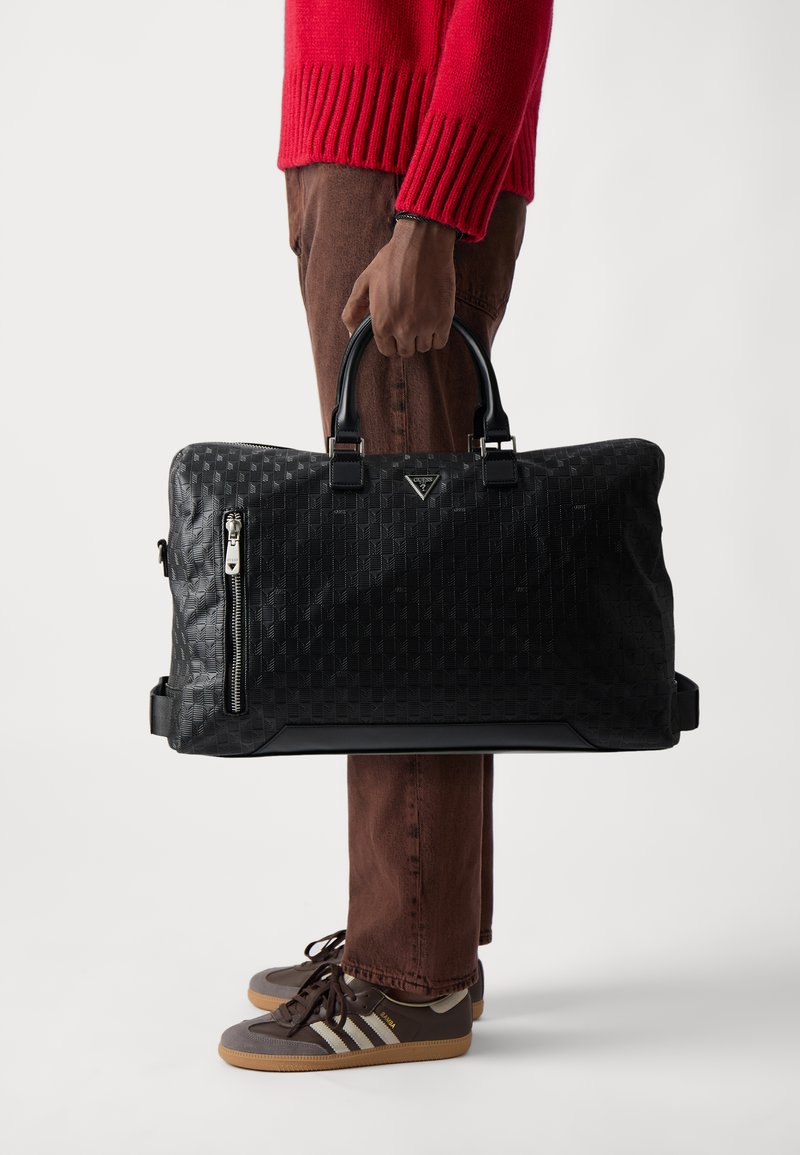Bolso de viaje negro con un patrón texturizado, cremallera lateral y asas planas, hecho de material sintético. Posicionado en posición vertical, sujeto por una persona.
