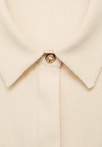 Beige overhemd met kraag van een gestructureerde stof, met een enkele knoopsluiting. De kraag is plat en het materiaal heeft een subtiele glans.