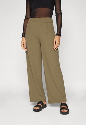 Brede cargobroek van olijfgroen stof met twee voorzakken, gecombineerd met een doorschijnende zwarte blouse met lange mouwen en zwarte sandalen met straps.