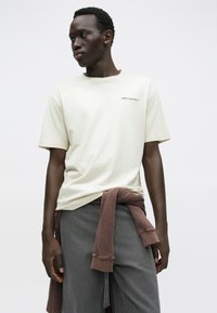 Bézs kokvilnas T-krekls ar īsām piedurknēm un oranžu McLaren logotipu. Kombinēts ar pelēkām biksēm un brūnu džemperi, kas sasiets ap vidukli.