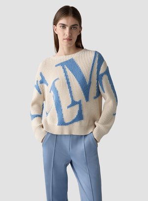 Junge Frau trägt einen cremefarbenen Strickpullover mit großen blauen Buchstaben und hat die Hände in den Taschen einer hellblauen Hose.