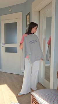 Šedá mikina s nápisem "Wellness Club", kterou drží osoba v světle růžovém topu, bílých kalhotách s rozšířenými nohavicemi a modrých botách. Viditelné zrcadla a dřevěná podlaha.