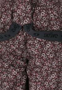 Brązowe spodnie w kwiatowy wzór z różowymi i białymi kwiatami; mają czarną elastyczną talię z nadrukowanym logotypem. Faktura wydaje się miękka i lekka.