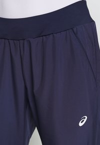 Tmavě modré sportovní kraťasy vyrobené z lehké látky. Mají pružný pas a bílý logo na dolní levé straně. Texturovaný povrch.