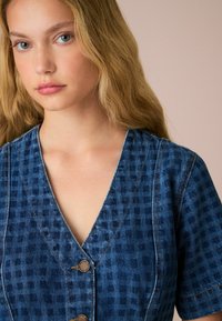 Abito in denim con maniche corte, scollo a V profondo e dettagli con bottoni. Presenta un motivo a quadrettoni blu scuro e dettagli di cuciture a contrasto.