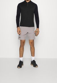 Man som bär en svart långärmad tröja, ljusgråa shorts, vita strumpor och svarta sneakers, håller en brun vattenflaska i höger hand.