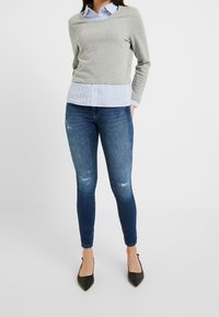 Maglione grigio sopra una camicia azzurra a righe, abbinato a jeans skinny blu scuro con dettagli consumati e ballerine nere a punta.