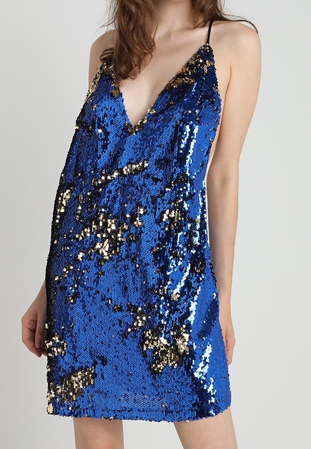 Mini-robe bleue sans manches avec motif en sequins dorés et noirs et fines bretelles, portée par une femme aux cheveux lâchés.