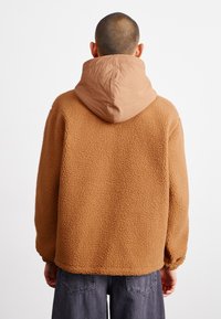 Casaco de lã castanho com superfície texturizada, apresentando um capuz em tecido bege e um design solto. Punhos elegantes e barra caída.