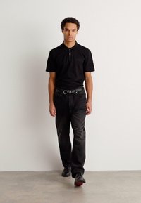 Črna polo majica s ovratnikom, v kombinaciji s temnimi kavbojkami in črnimi čevlji. Model v nevtralnem stoječem položaju na enobarvni podlagi.