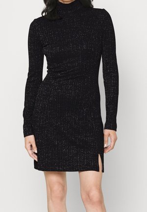 Femme portant une robe en maille côtelée noire à manches longues, col montant et une petite fente latérale au-dessus du genou.