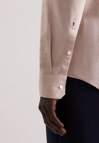 Camicia button-up beige chiaro con polsino che presenta un bottone bianco e un dettaglio ricamato nero. Tessuto morbido con vestibilità su misura.