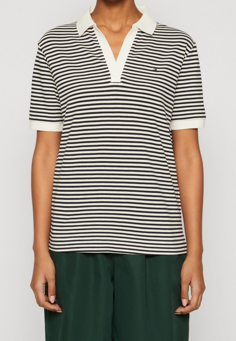 Personne portant un polo à rayures horizontales noires et blanches, manches courtes, avec un col blanc et un pantalon vert foncé.