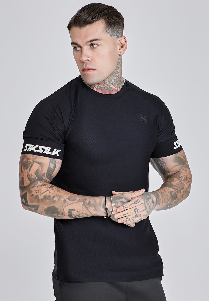 Fekete, testhez simuló póló rövid ujjakkal, texturált anyaggal. Az "SIKSILK" logó fehér szalag formájában a bal kar körül. Egyéb részletek között látható tetoválások is szerepelnek.