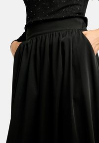 Melna svārki ar pieguļošu jostasvietu un kroku dizainu. Izgatavoti no gludas auduma, ar sānu kabatām un plūstošu siluetu.