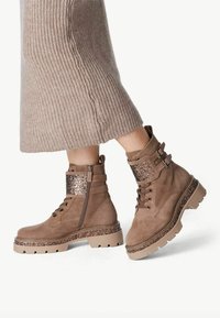 Bottines en suède marron avec une bande décorative en paillettes, fermeture éclair sur le côté et semelle robuste. Agrémentées d'éléments beige clair et métalliques.