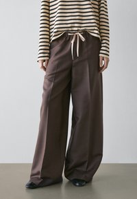 Persona che indossa pantaloni marroni a gamba larga con vita con coulisse, scarpe nere e una maglia a maniche lunghe a righe beige e nere su sfondo semplice.