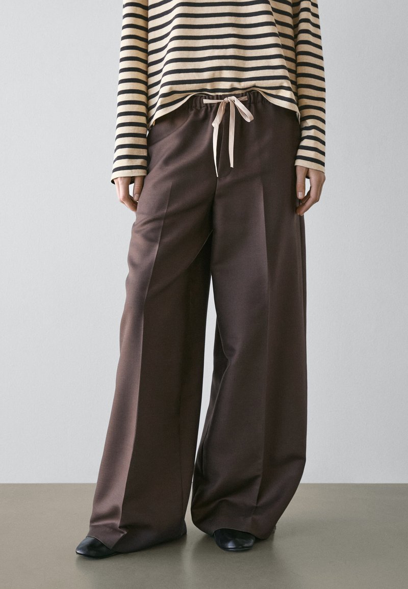 Persona che indossa pantaloni marroni a gamba larga con vita con coulisse, scarpe nere e una maglia a maniche lunghe a righe beige e nere su sfondo semplice.